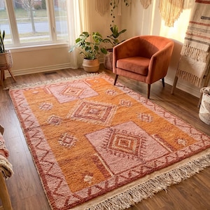 May include: A geometric patterned rug in shades of orange and pink, with a fringed edge. The rug's design includes diamond and square shapes. It is placed on a wooden floor in a room with natural light.