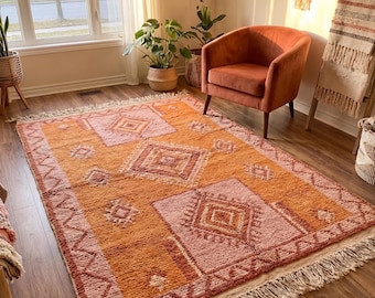 May include: An orange and pink geometric patterned rug with a fringed edge. The rug features diamond and square shapes in varying shades of orange and pink. The rug is placed on a wooden floor in a well-lit room.