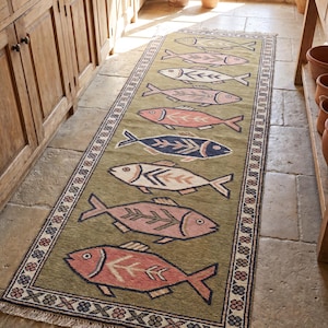 Könnte beinhalten: Ein rechteckiger, olivgrüner Teppich mit einem sich wiederholenden Fischmuster in Rosa, Weiß und Marineblau. Der Teppich hat einen dekorativen Rand mit floralen und geometrischen Elementen. Er liegt auf einem Steinboden.