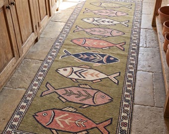 May include: A rectangular olive green rug with a repeating fish pattern in shades of pink, white, and navy blue. The rug has a decorative border with floral and geometric elements. The rug is placed on a stone floor.