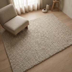 Puede incluir: Una alfombra de área texturizada de tonos neutros con una superficie densa y nudosa. La alfombra es rectangular y cubre una porción de un suelo de madera clara. Un sillón beige se encuentra sobre la alfombra, cerca de una ventana con cortinas transparentes.