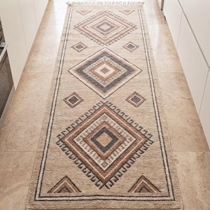 Op de afbeelding: Een lang rechthoekig loperkleed met een beige basis en een herhalend patroon van grote ruiten in tinten bruin, antraciet en crème. Het kleed heeft een getextureerde pool, een donkere rand en decoratieve franjes aan beide korte zijden.