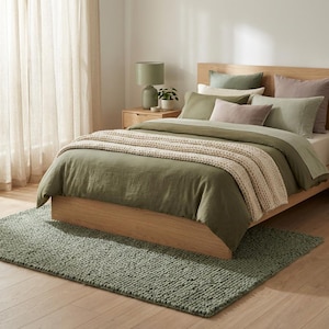 May include: A bedroom scene featuring a bed with olive green bedding, including a duvet cover and pillows. A beige knitted throw is draped over the bed. A sage green rug sits on the wooden floor. A wooden nightstand with a lamp and plant is visible.