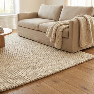 K&ouml;nnte beinhalten: Ein Wohnzimmer mit beigem Sofa und cremefarbener Decke. Ein runder Holztisch mit B&uuml;chern. Ein strukturierter, cremefarbener Teppich liegt auf einem Holzboden. Der Raum hat eine neutrale Farbpalette und wirkt einladend.