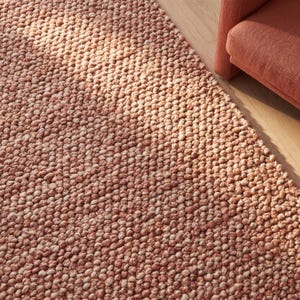 Puede incluir: Una alfombra texturizada con nudos redondos en tonos melocotón y crema. La superficie de la alfombra está densamente cubierta con pequeños nudos en relieve, creando un patrón táctil y visualmente interesante. La alfombra está sobre un suelo de madera, con un sillón melocotón.