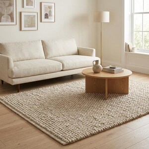 May include: A living room scene featuring a beige sofa, a round wooden coffee table, and a textured beige rug. The room is decorated with framed artwork and a floor lamp, creating a cozy and inviting atmosphere.