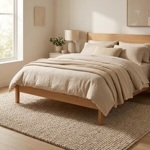 May include: A bedroom scene featuring a bed with beige bedding, a knitted throw, and pillows. The bed frame is made of light-colored wood. A beige rug covers the floor. A lamp and artwork are also visible.