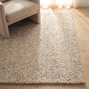 Puede incluir: Una alfombra beige texturizada con un pelo nudoso y en bucle. La alfombra es rectangular y se asienta sobre un suelo de madera clara. Una butaca beige es parcialmente visible en la esquina superior izquierda, con luz solar entrando por una ventana.