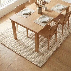 K&ouml;nnte beinhalten: Ein Esstisch aus hellem Holz mit vier passenden St&uuml;hlen, gedeckt f&uuml;r eine Mahlzeit. Der Tisch steht auf einem cremefarbenen, strukturierten Teppich. Der Raum hat einen hellen Holzboden und ein Fenster im Hintergrund. Der Tisch ist mit Tellern und Tischsets gedeckt.