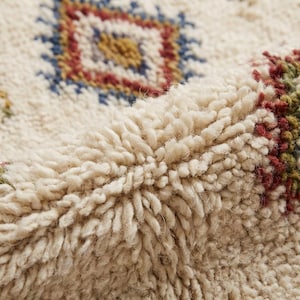May include: Close-up of a cream-colored, textured rug with a geometric pattern. The design features a central diamond shape in blue, red, and yellow. The rug has a thick, plush pile, suggesting a soft and comfortable texture.