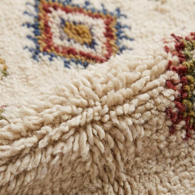May include: Close-up of a cream-colored, textured rug with a geometric pattern. The design features a central diamond shape in blue, red, and yellow. The rug has a thick, plush pile, suggesting a soft and comfortable texture.