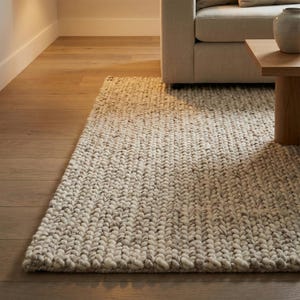 Puede incluir: Una alfombra rectangular con una textura de tejido grueso en una mezcla de hilos color crema, beige y gris claro. La alfombra está construida con grandes bucles de hilo visibles, creando una superficie densa y táctil que cubre el suelo.