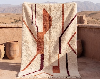 May include: Cream-colored rug with an abstract geometric design in shades of brown and burgundy. The rug has a textured surface and a fringe along the bottom edge. The rug is displayed outdoors.