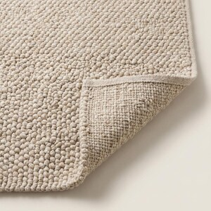 May include: A beige, textured bath mat with a looped pile. The corner of the mat is folded back, revealing a woven underside. The mat has a neutral color and a simple design, suitable for a bathroom.