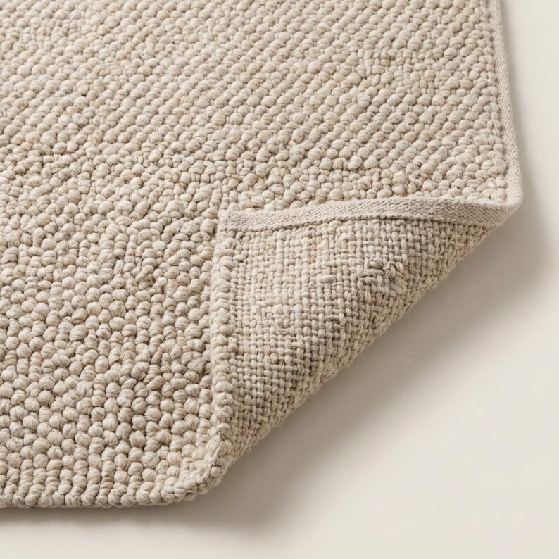 May include: A beige, textured bath mat with a looped pile. The corner of the mat is folded back, revealing a woven underside. The mat has a neutral color and a simple design, suitable for a bathroom.