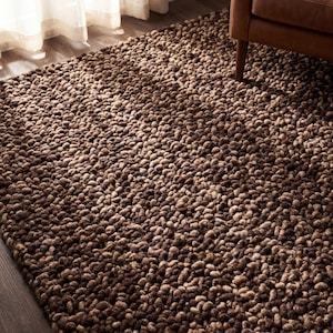 May include: A brown, textured rug with a dense, looped pile. The rug has varying shades of brown, creating a rich, warm appearance. The rug is placed on a wooden floor, with a glimpse of a brown leather sofa leg in the background.
