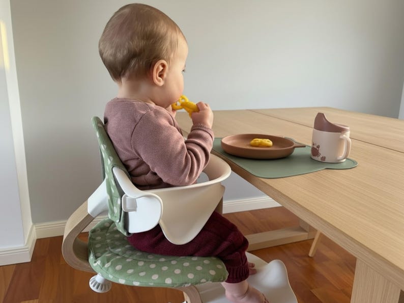 May include: A baby sits in a high chair with a white tray and a green polka dot seat. A wooden plate with food, a cup, and a placemat are on the table. The baby wears a pink shirt and burgundy pants.