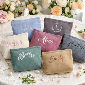 Puede incluir: Una colección de neceseres de pana personalizados en varios colores, incluyendo azul, rosa y beige. Cada neceser está bordado con un nombre o inicial. Los neceseres están dispuestos sobre un mantel de encaje blanco, con arreglos florales en el fondo.