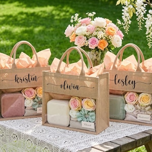 May include: Three burlap tote bags with clear front panels, each personalised with a name in black script. The bags are filled with gifts, decorative flowers, and tissue paper. A bouquet of flowers and a bottle of champagne are in the background.