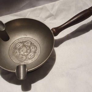 Op de afbeelding: Een vintage metalen asbak met een donkerhouten handvat. De asbak is rond met twee ingebouwde rustplaatsen en een bloemenontwerp in het midden. De handgreep is ongeveer 15 cm lang.
