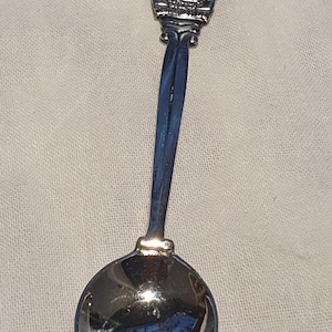 Op de afbeelding: Een zilveren souvenirlepel met een blauw geëmailleerd embleem aan de bovenkant. Het embleem toont een figuur en een decoratieve rand. De lepel heeft een lange, slanke steel en een afgeronde kom. De lepel is waarschijnlijk gemaakt van roestvrij staal.
