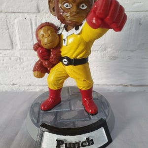 Op de afbeelding: Een beeldje van een aapachtig personage in een gele overall met rode laarzen en handschoenen, met een kleinere aap. De basis is grijs met het woord "Punch" op een zwart-wit plaatje.
