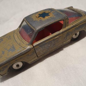 May include: A vintage toy car with a weathered, distressed appearance. The car is primarily olive green with a gray roof and red interior. A blue star emblem is visible on the roof. The toy car has white wheels.