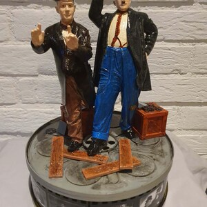 Peut inclure: Figurine détaillée de Laurel et Hardy, le duo comique, sur une base de bobine de film. Les personnages portent leurs tenues emblématiques, l'un d'eux tenant un chapeau melon. La base présente des détails de bande de film et des accents en bois.