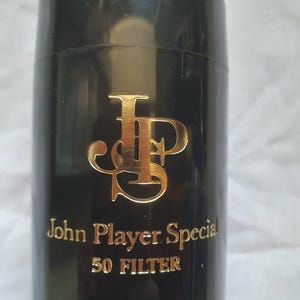 May include: A black cylindrical container with gold lettering and logo. The text "John Player Special" and "50 FILTER" are visible. The container has a glossy finish.
