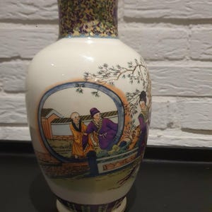 Op de afbeelding: Een witte keramische vaas met een bolvormige body en een uitlopende rand. De vaas heeft een gedetailleerde geschilderde scène van figuren in traditionele kleding in een cirkelvormig kader. De hals heeft een decoratief patroon in goud, paars en groen.