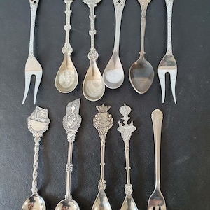 Puede incluir: Una colección de cucharas y tenedores de plata antiguos. Las cucharas presentan diseños decorativos variados, como un velero, un edificio y una figura. Los tenedores tienen mangos ornamentados. Los artículos están dispuestos sobre una superficie oscura.