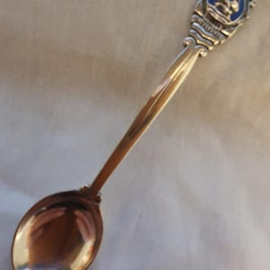 Op de afbeelding: Een zilveren lepel met een gedetailleerde handgreep en een blauw emaille embleem. De lepel heeft een lange, slanke steel en een afgeronde kom. Het embleem heeft een ontwerp met een blauwe achtergrond en zilveren accenten.