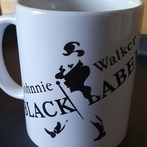 Op de afbeelding: Witte keramische mok met het Johnnie Walker Black Label logo. Het logo toont een zwart silhouet van een man die loopt, met de merknaam in zwarte letters. De mok heeft een gebogen handvat en een glad oppervlak.