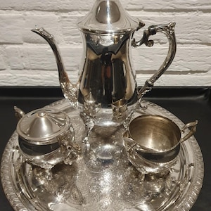 May include: A silver tea set on a silver tray. The set includes a teapot, a sugar bowl, and a creamer. The tray and set have ornate detailing. The teapot has a decorative handle and a lid with a finial.