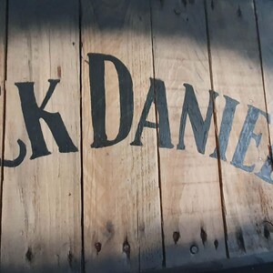 Op de afbeelding: Houten bord met de tekst "Jack Daniel's" in zwarte en blauwe letters. Het bord is gemaakt van houten planken met zichtbare nerven en knopen. De letters zijn licht verweerd, wat het een vintage uitstraling geeft.