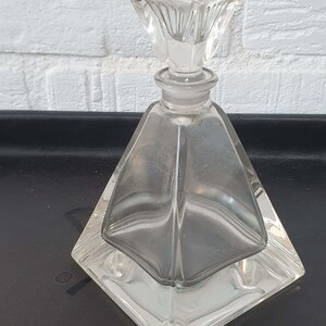 May include: A clear glass perfume bottle with a square base and a pyramid-shaped body. The bottle has a square stopper. The design is vintage and elegant, suitable for display or use. The bottle is made of clear glass.