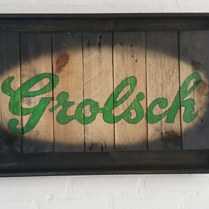 Op de afbeelding: Houten bord met het woord "Grolsch" in groene, gestileerde letters. Het bord is geplaatst in een donkere lijst en gemonteerd op een witte bakstenen muur. Het hout heeft een natuurlijke, lichtbruine kleur.
