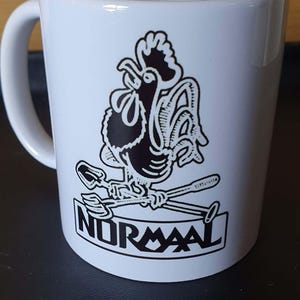 Op de afbeelding: Witte keramische mok met een zwarte haan illustratie, gekruiste lepels en het woord "NORMAAL". De mok heeft een gebogen handvat en een glanzende afwerking.