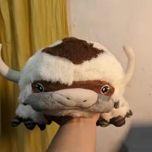 May include: A plush toy resembling a flying bison, featuring a white body with a brown patch on its head and back. It has large, expressive eyes, small horns, and four feet. The toy is held up by a person's hand.
