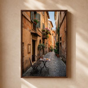 以下が含まれることがあります： イタリアの町の狭い石畳の路地を描いた額装されたウォールアートプリント。温かみのある黄土色の建物、緑の鎧戸、花で飾られたバルコニー、そして通りに置かれた2脚の椅子がある小さなビストロテーブルが特徴的な風景です。