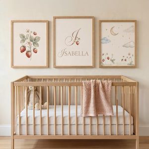 Op de afbeelding: Drie ingelijste kunstprints voor de kinderkamer boven een houten wieg. De prints tonen aquarel illustraties van aardbeien, een monogram met de naam "Isabella" en een nachtscène met een maan en wolken. Een roze deken is over de wieg gedrapeerd.