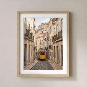 K&ouml;nnte beinhalten: Ein gerahmter Druck einer engen europ&auml;ischen Stra&szlig;e mit einer gelben Stra&szlig;enbahn auf Gleisen. Geb&auml;ude mit Balkonen und verschiedenen Pastellfarben s&auml;umen die Kopfsteinpflasterstra&szlig;e. Der Druck ist in einem hellen Holzrahmen.