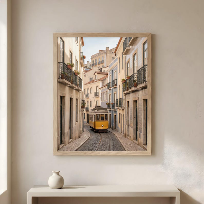 K&ouml;nnte beinhalten: Gerahmter Druck einer Stra&szlig;enansicht von Lissabon. Eine gelbe Stra&szlig;enbahn f&auml;hrt eine Kopfsteinpflasterstra&szlig;e entlang, die von Geb&auml;uden in Beige-, Blau- und Wei&szlig;t&ouml;nen ges&auml;umt ist. Das Bild ist in einem hellen Holzrahmen gerahmt und weckt ein Gef&uuml;hl von Reise und europ&auml;ischem Charme.