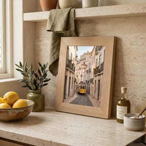 K&ouml;nnte beinhalten: Ein gerahmtes Foto einer europ&auml;ischen Stra&szlig;enszene mit einer gelben Stra&szlig;enbahn auf Schienen. Der Rahmen ist aus hellem Holz. Eine Sch&uuml;ssel mit Zitronen, eine Vase mit Gr&uuml;n und eine Flasche Oliven&ouml;l sind ebenfalls zu sehen.