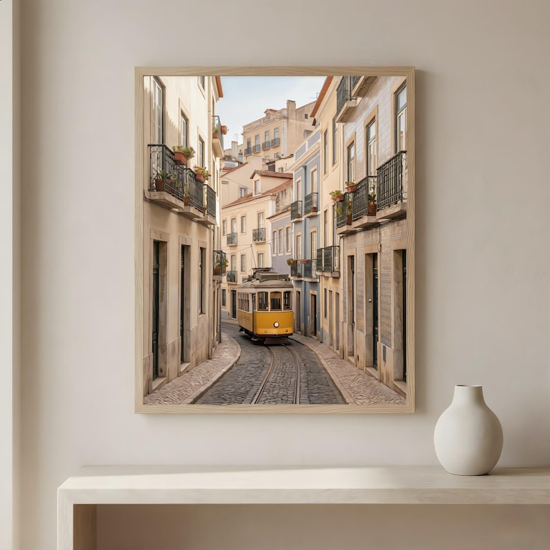 K&ouml;nnte beinhalten: Gerahmter Druck einer Stra&szlig;enszene in Lissabon, Portugal. Eine gelbe Stra&szlig;enbahn f&auml;hrt auf den Gleisen in der Mitte der Kopfsteinpflasterstra&szlig;e. Geb&auml;ude mit Balkonen und farbenfrohen Fassaden s&auml;umen die Stra&szlig;e. Der Druck ist in einem hellen Holzrahmen.