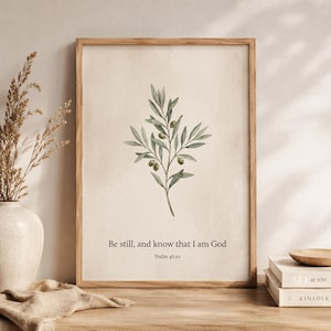 May include: A framed print featuring a watercolor illustration of an olive branch with green leaves and olives. The text "Be still, and know that I am God, Psalm 46:10" is printed below the illustration. The frame is a light brown color.