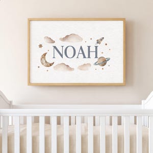 Può includere: Una stampa incorniciata per la cameretta, con il nome "NOAH" in blu, circondato da illustrazioni ad acquerello di nuvole, una luna crescente, stelle, un razzo e un pianeta. L'opera è su uno sfondo bianco strutturato.