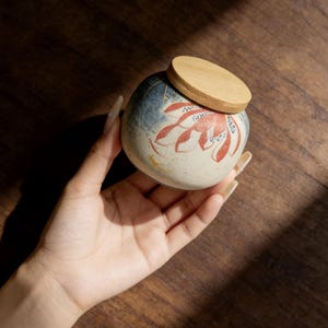 May include: A small, round ceramic jar with a wooden lid. The jar features a floral design in red and white against a cream background. The top of the jar has a blue and white design. The lid is made of light-coloured wood.