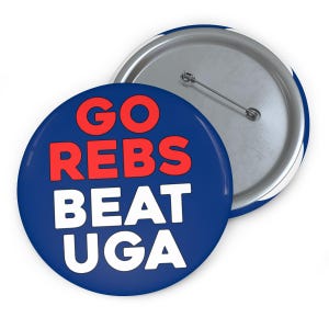 Könnte beinhalten: Ein runder Ansteckbutton mit dunkelblauem Hintergrund. Der Text GO REBS ist in fetten roten Buchstaben in den oberen zwei Zeilen gedruckt, und der Text BEAT UGA ist in fetten weißen Buchstaben in den unteren zwei Zeilen gedruckt. Eine Nadel ist sichtbar.