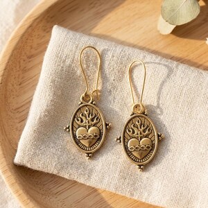 May include: A pair of gold-colored earrings with an oval pendant featuring a heart, cross, and flames design. The earrings are displayed on a beige cloth, with a wooden tray in the background.